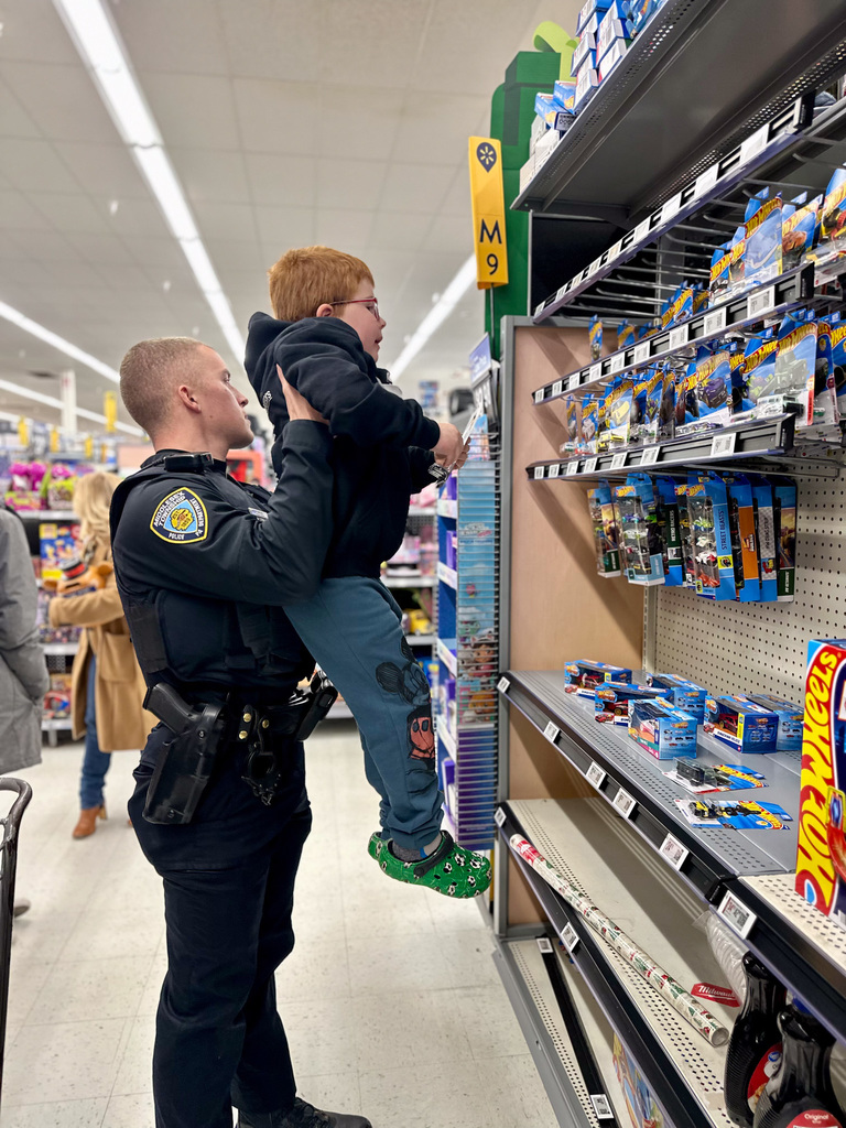 A police officer lifts a child to reach toy items on a store shelf during the 11th annual Shop with a Cop event.