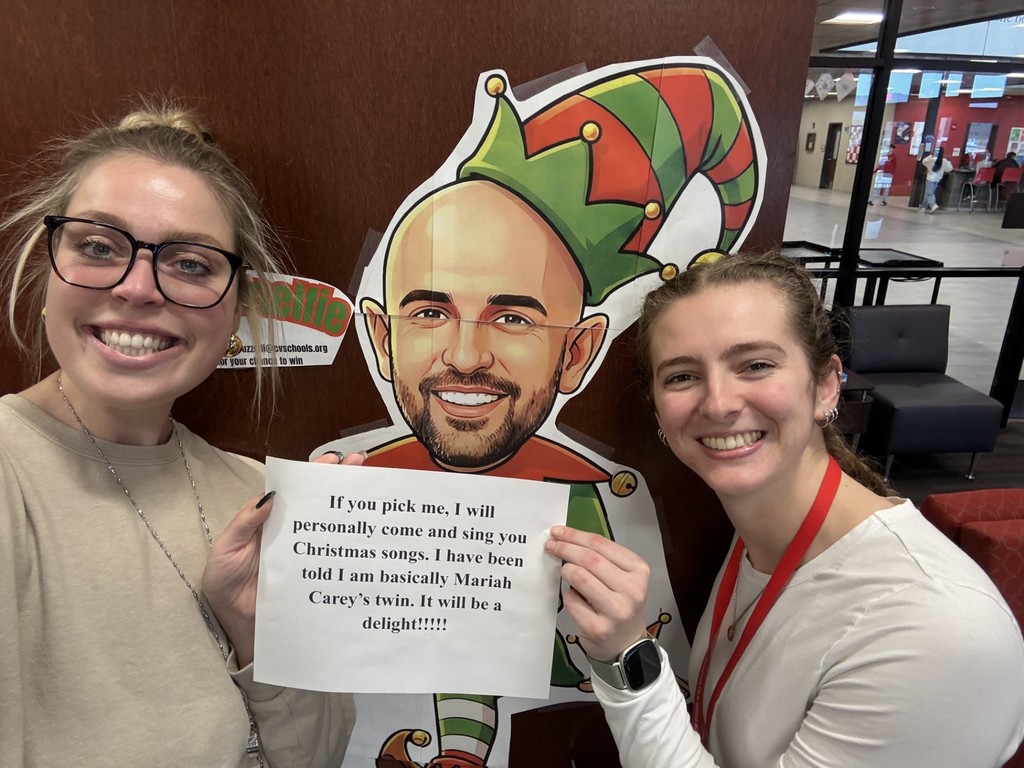 Two staff members at Mountain View Middle School take a selfie with a cardboard elf named Snelfie, holding a humorous sign about singing Christmas songs.