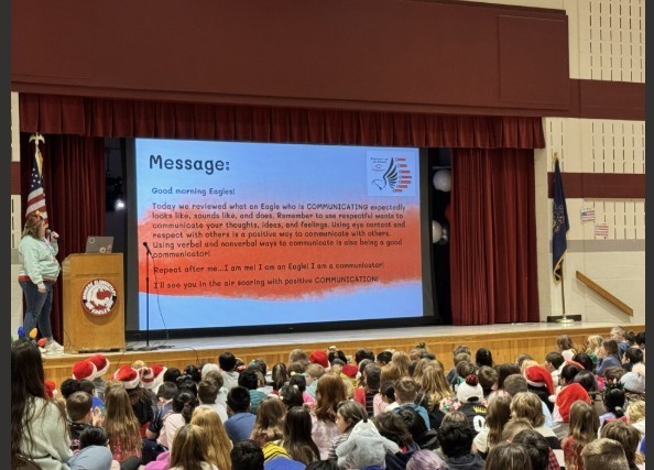 Teacher standing on stage reading the morning message to students.