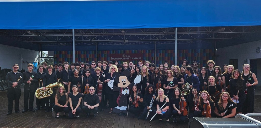 The Cumberland Valley orchestra poses together with their instruments on a Disney Imagination Campus stage, joined by a cutout of Mickey Mouse.