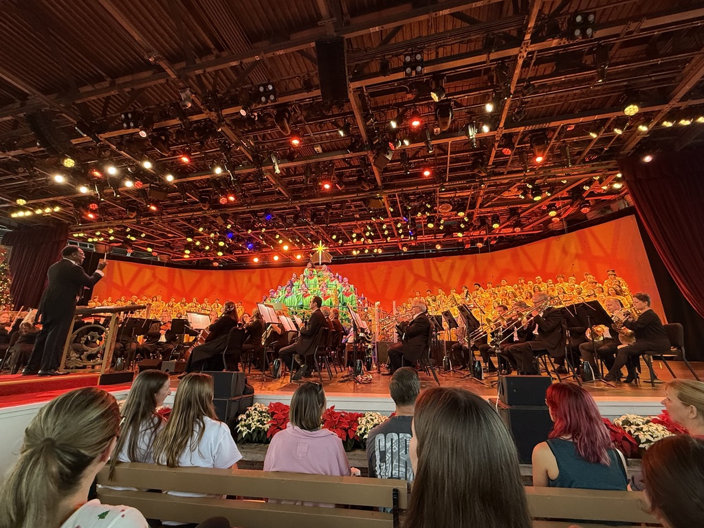 Cumberland Valley Choirs perform in EPCOT’s Candlelight Processional with a full orchestra and a brightly lit Christmas tree backdrop, part of a program narrated by celebrity Jordan Fisher.