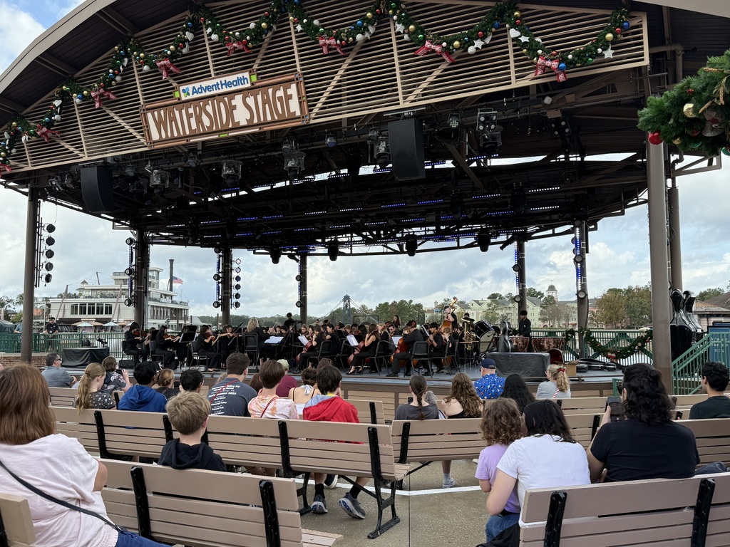 The Cumberland Valley orchestra performs on Disney’s Waterside Stage, playing for a seated outdoor audience beneath holiday decorations.