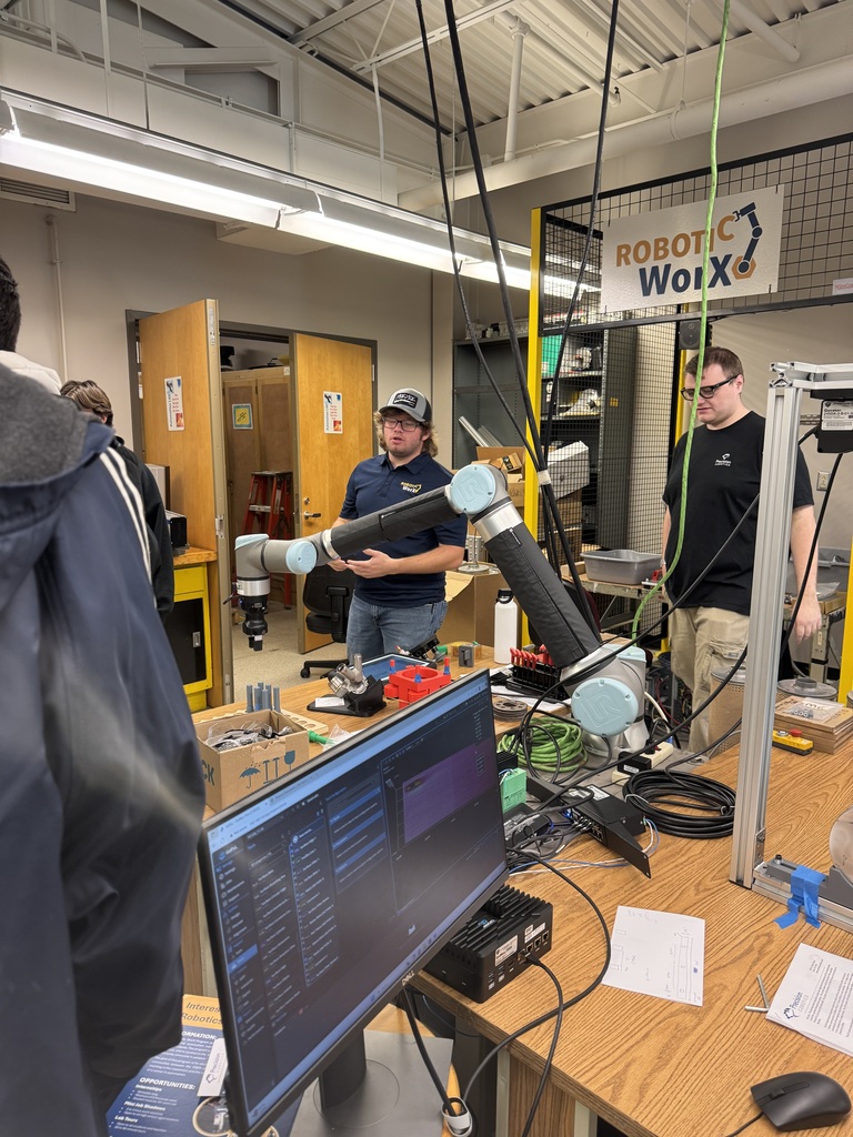 CVHS students observe a robotics demonstration at Millersville University during AEST Exploration Day, with robotic arms and computer equipment on a workbench.