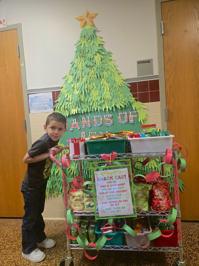 Student standing next to a festive snack cart decorated with red and green paper chains and filled with assorted snacks and drinks. Behind the cart is a large handmade Christmas tree display with a star on top and the words 'Hands of Joy' in colorful letters.