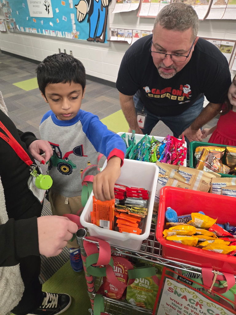 Student selects a snack from a festive cart decorated with red and green paper chains, filled with colorful treats like candy bars and chips. Adults stand nearby assisting, and the background shows a classroom wall with educational displays.