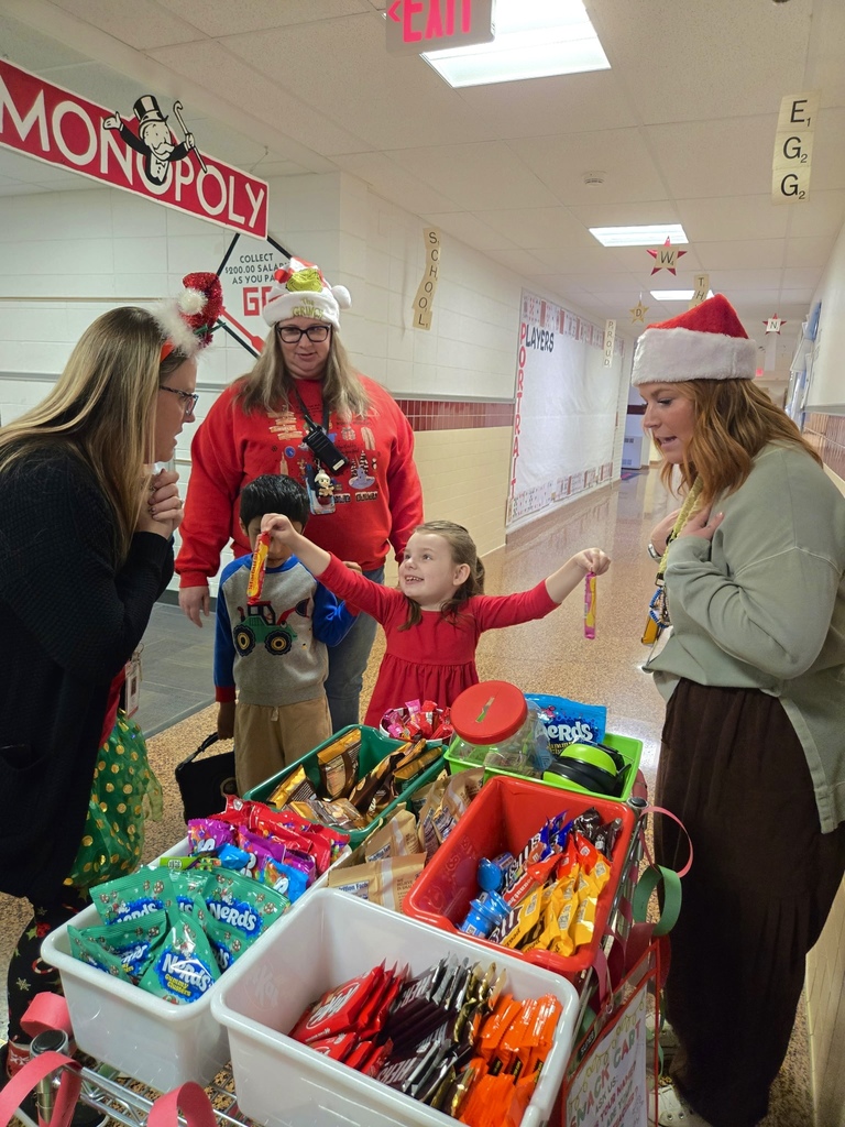Monroe Elementary hallway scene with a festive snack cart filled with colorful treats like chips, candy, and drinks. Two children are handing out snacks to staff members wearing holiday-themed attire and accessories.
