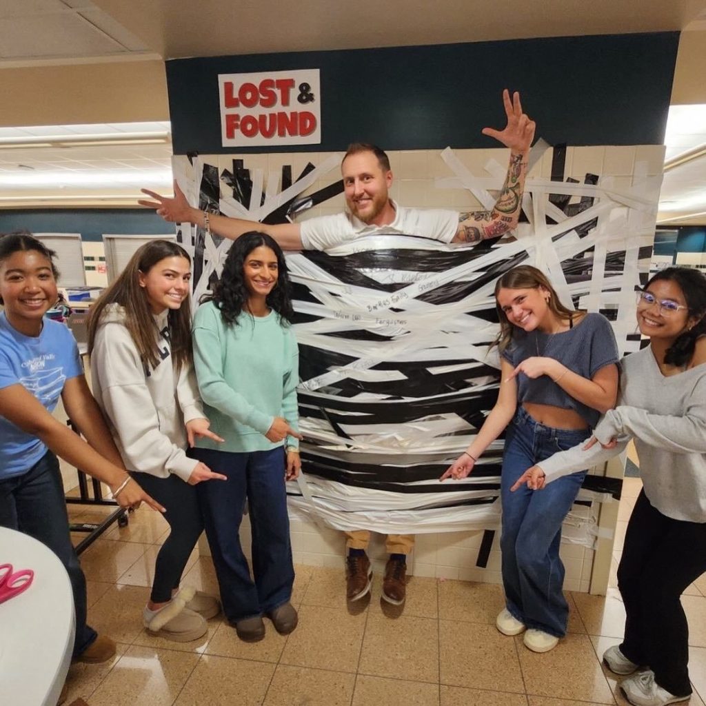 Mr. Shaw, CVHS Dean, is taped to a wall with layers of black and white tape while students stand around pointing and smiling during a CV Mini-THON fundraiser