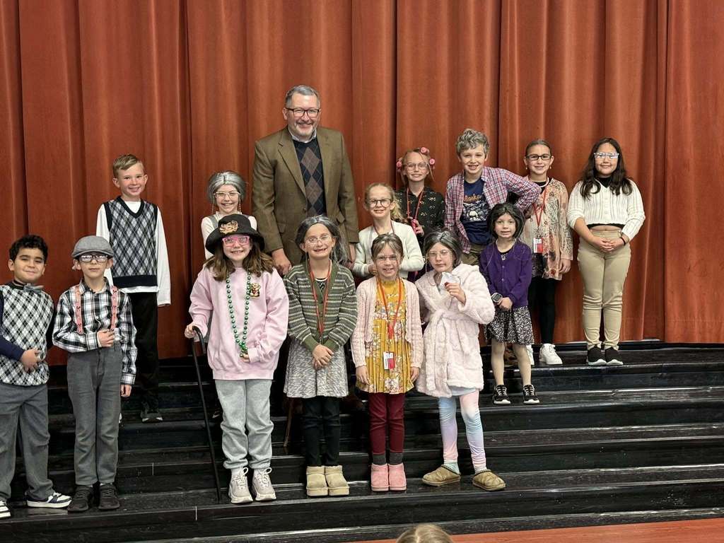 A group of students and their Principal dressed as elderly adults stand on risers to celebrate the 100th day of school.