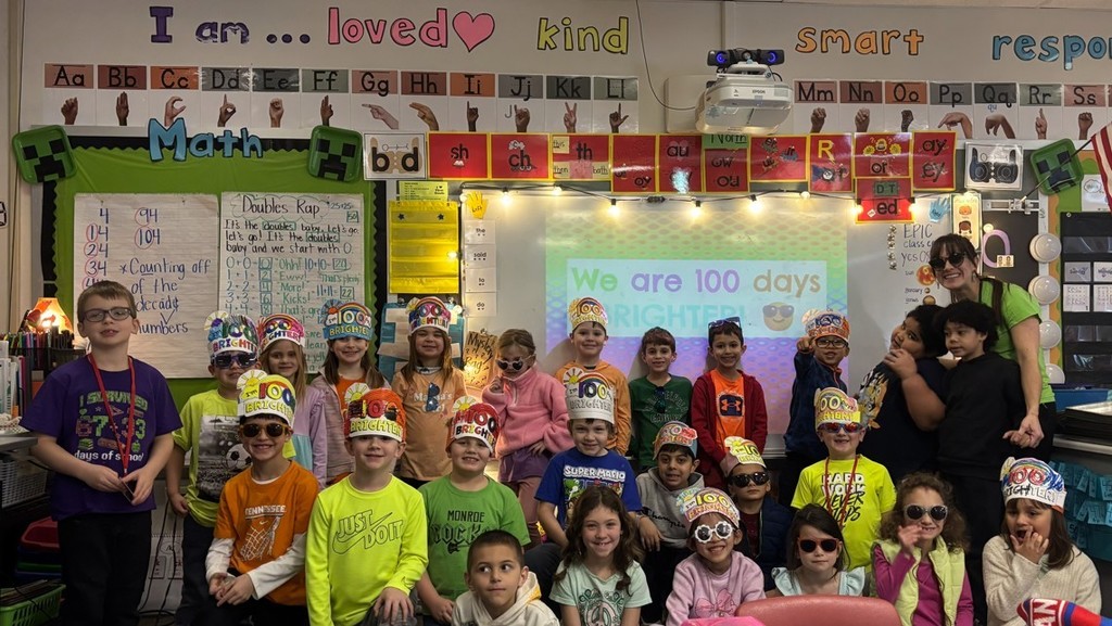 Elementary school class lined up and smiling with teacher, wearing hand-made paper hats that say 100 days brighter
