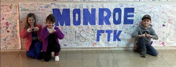 Three students kneeling in front of sign that says Monroe is FTK in blue letters.