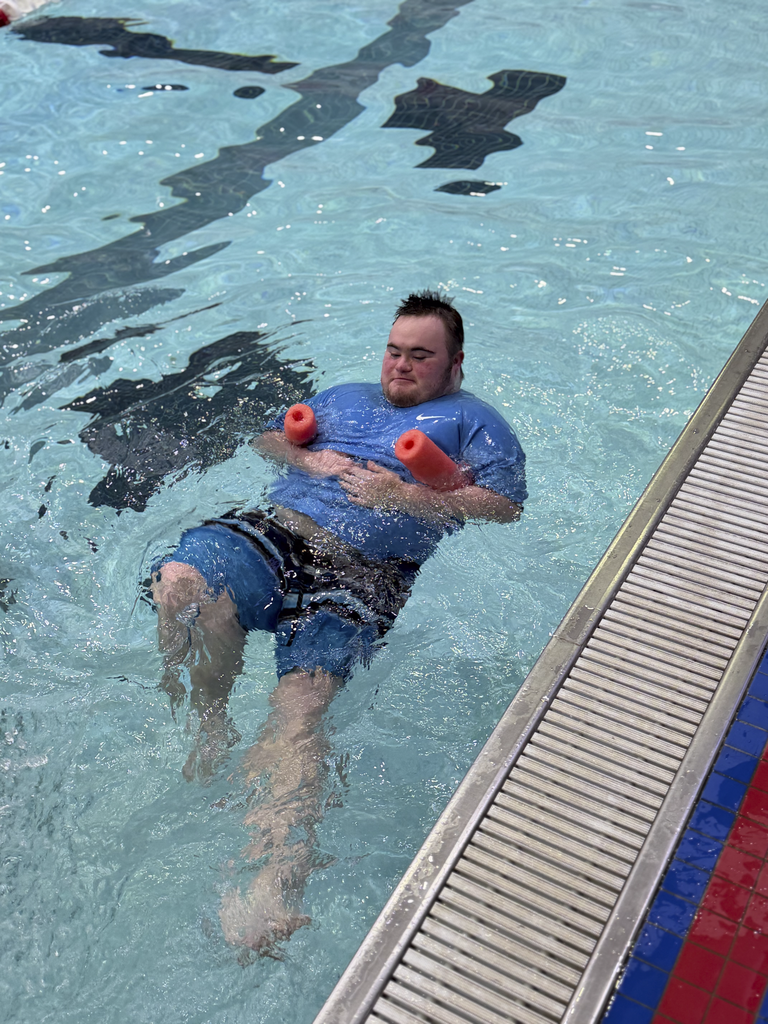 A swimmer floating on their back in an indoor pool while holding onto a foam noodle during a water exercise.