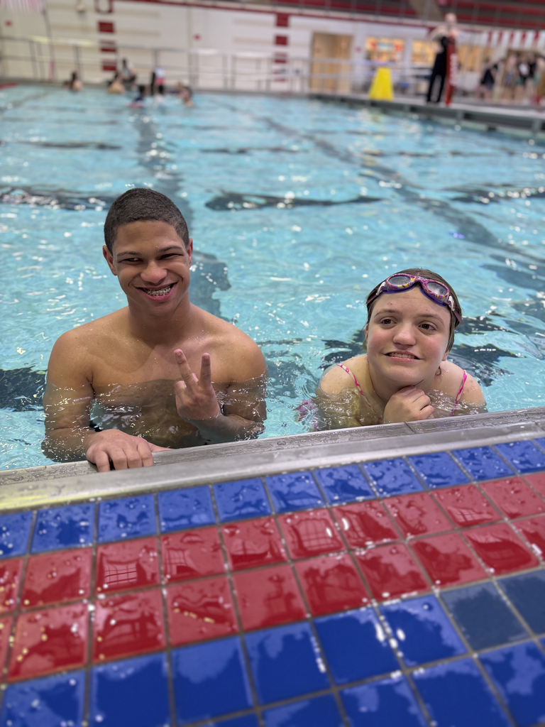 Two people at the edge of an indoor swimming pool, leaning on the tiled deck, with one person holding up the peace sign.