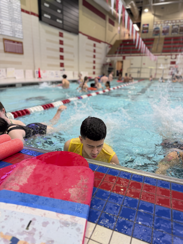A swimmer at the pool edge practicing a kicking drill while holding the wall, with other swimmers in the background.