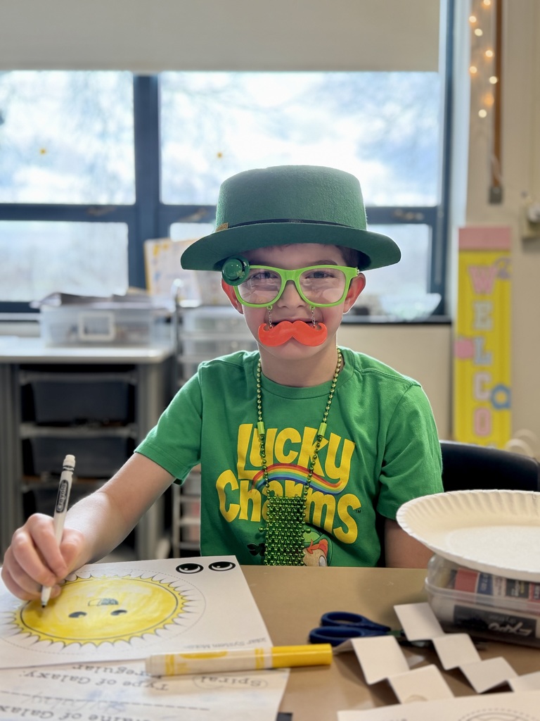Student dressed up as a leprechaun on St. Patrick's Day. He's sitting at a table with colored markers and smiling.