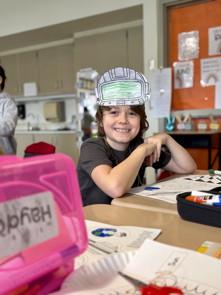 A third-grade student wears a paper astronaut helmet while sitting at a desk with space-themed worksheets during Monroe Elementary’s astronomy unit.
