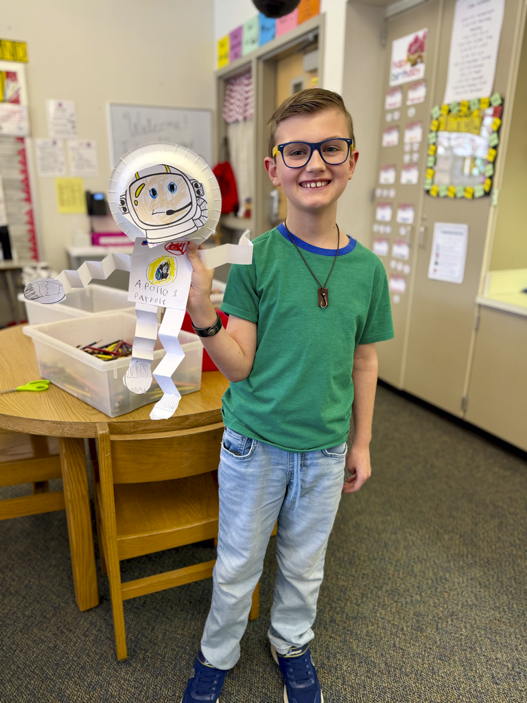 A third-grade student holds a paper astronaut craft during Monroe Elementary’s astronomy unit, standing in a classroom beside a table of art supplies.