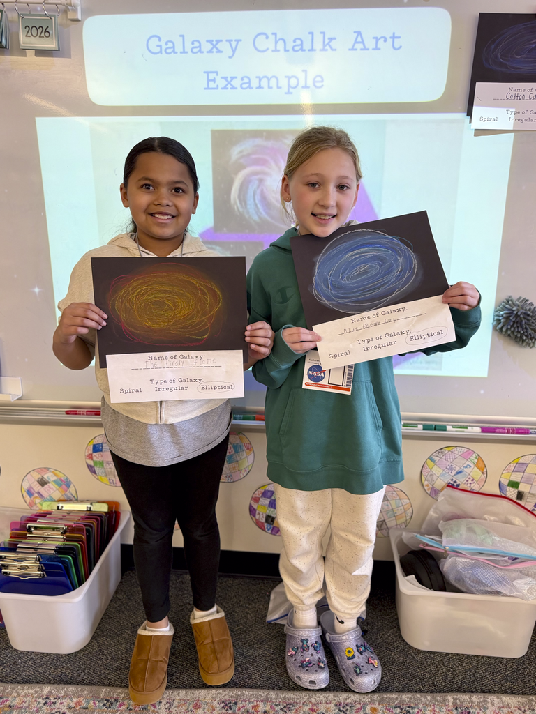 Two third-grade students display galaxy chalk art projects during an astronomy lesson, standing in front of a classroom whiteboard labeled “Galaxy Chalk Art Example.”