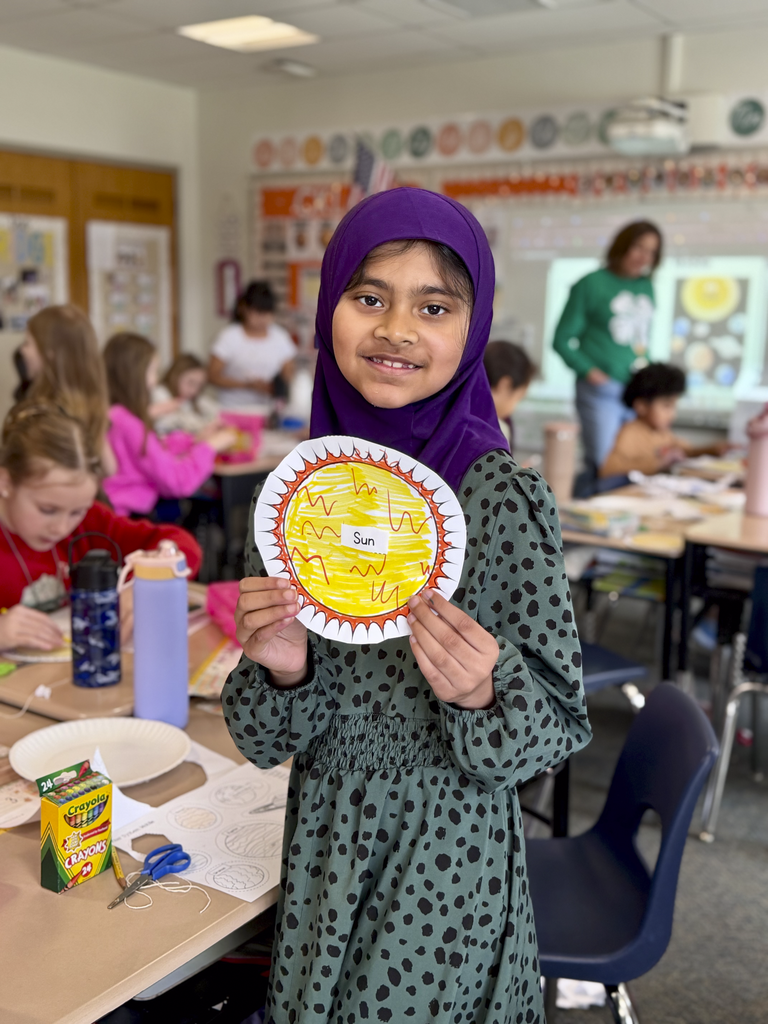 A third-grade student holds a paper sun artwork during Monroe Elementary’s astronomy unit, with classmates working on projects in the classroom behind them.