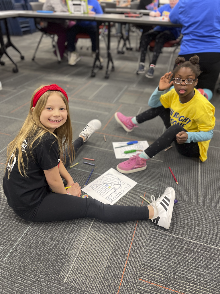 Two students are sitting on the floor, smiling, with coloring paper and colored pencils around them.