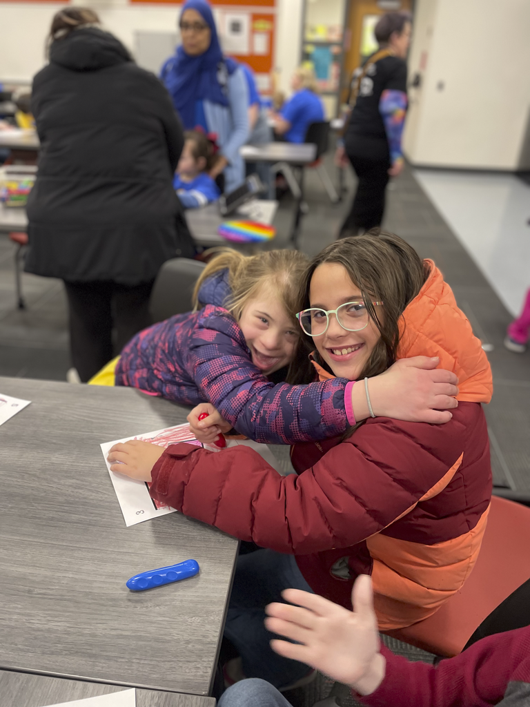 Two students share a hug at a table while coloring and smiling.