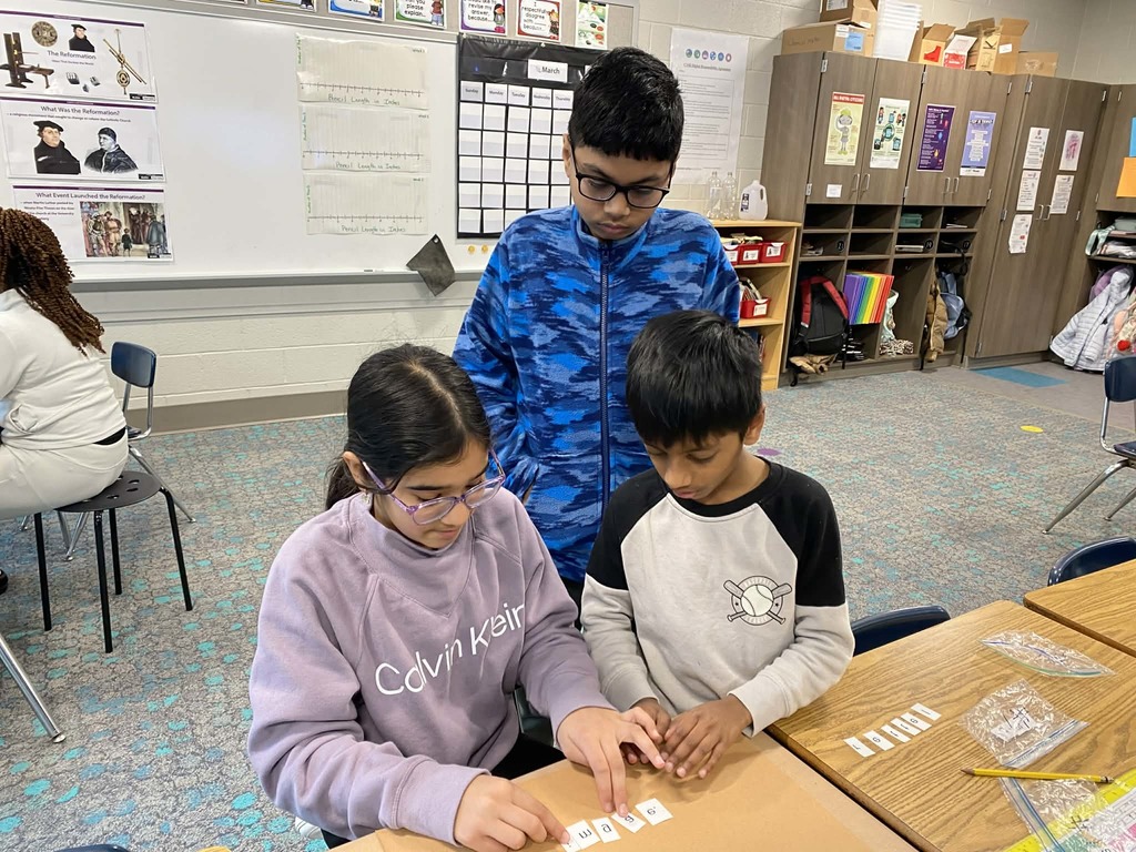  Fifth grade students arrange letter tiles together at a classroom table during a printing press lesson.