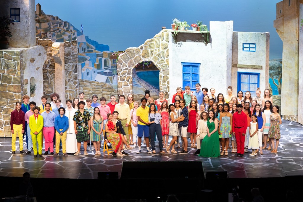 Full cast of CVHS’ Spring musical Mamma Mia! posed onstage in front of a Greek-style village set.