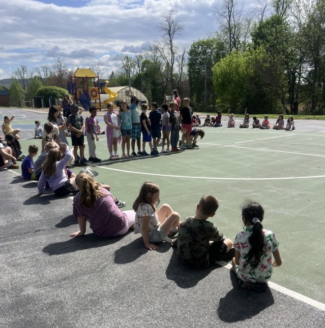 Students sitting on blacktop, some standing in a line.
