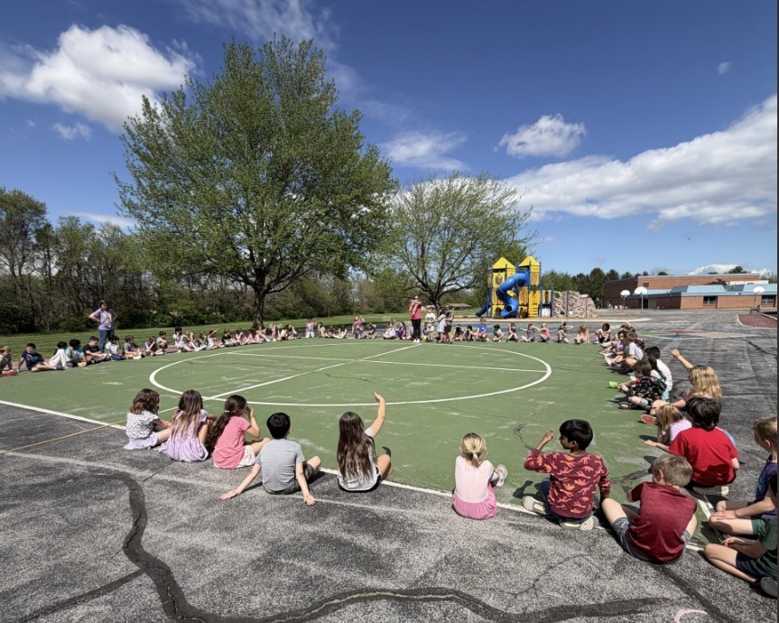 Students and teachers outside under the blue sky with trees filling up with green leaves.
