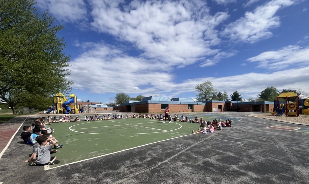 Students sitting on blacktop, listening to a teacher talk.