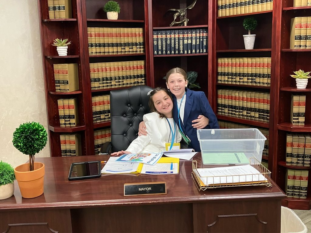 Two 5th grade students sit together at a mayor’s desk during a JA BizTown visit, with bookshelves behind them.