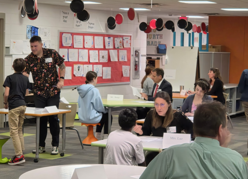 Adults interviewing students, sitting at tables in large room.
