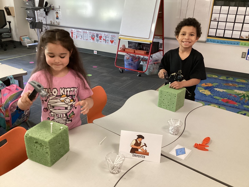 Two kindergarten students in a classroom hammer golf tees into green foam blocks during a hands-on activity.