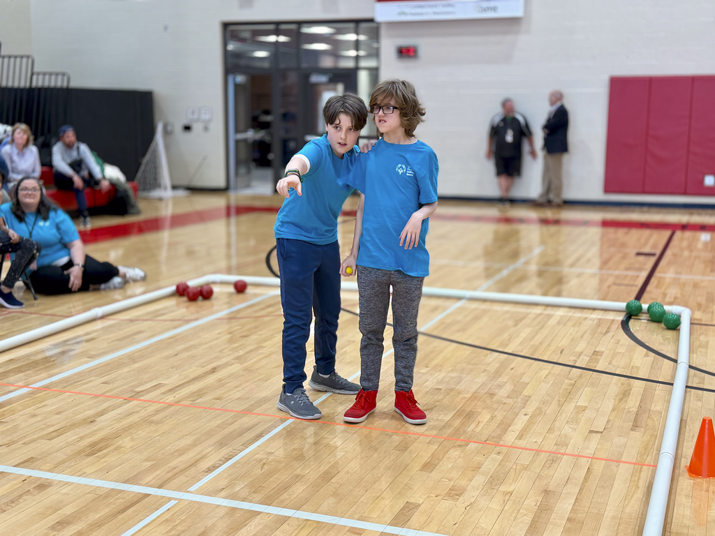 Two middle school students stand inside a gym bocce court during a unified bocce game, with one pointing while the other holds a ball.
