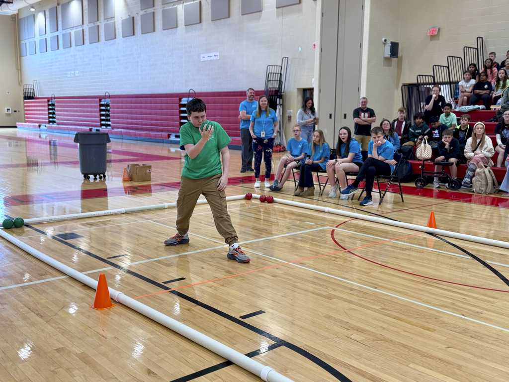 Middle school student winds up to throw a bocce ball inside a gym as peers and spectators watch from the sidelines.