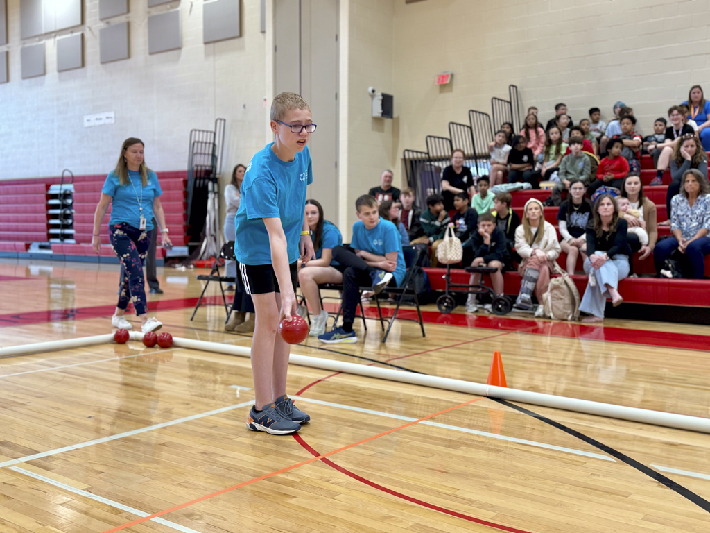 Middle school student rolls a red bocce ball during a unified bocce game in a gymnasium, with classmates and spectators watching from the bleachers.