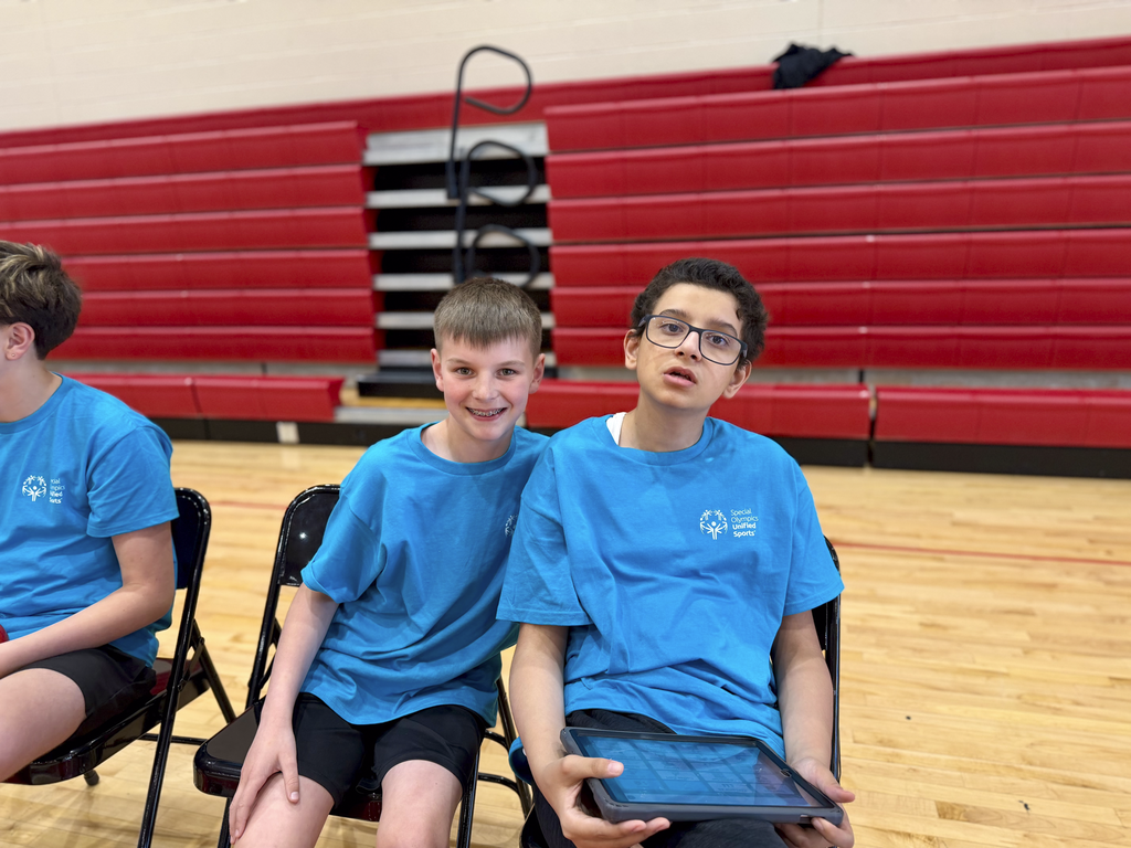 Two middle school students in blue Unified Sports shirts sit courtside in a gym during a unified bocce game, holding a tablet and watching the action.