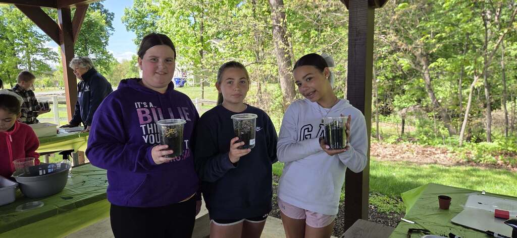 Three students hold clear cups with soil and plants under a pavilion during Middlesex Elementary’s 5th grade Environmental Ed Day.