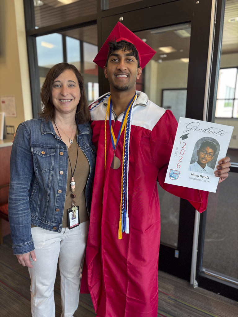 A student wearing a graduation cap and gown poses for a picture with their elementary teacher, hold a poster with their kindergarten picture on it.