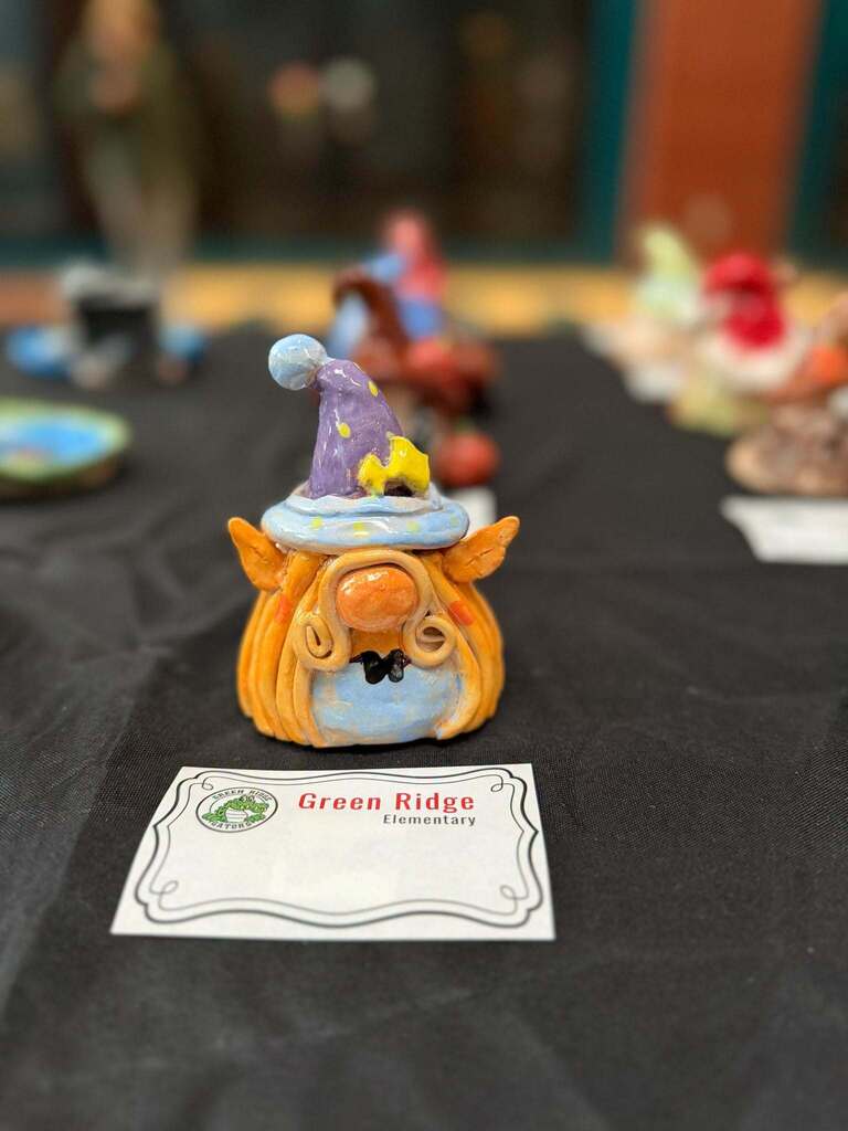 Small ceramic gnome figurine with a purple hat displayed on a table, labeled “Green Ridge Elementary,” with other student art blurred in the background.