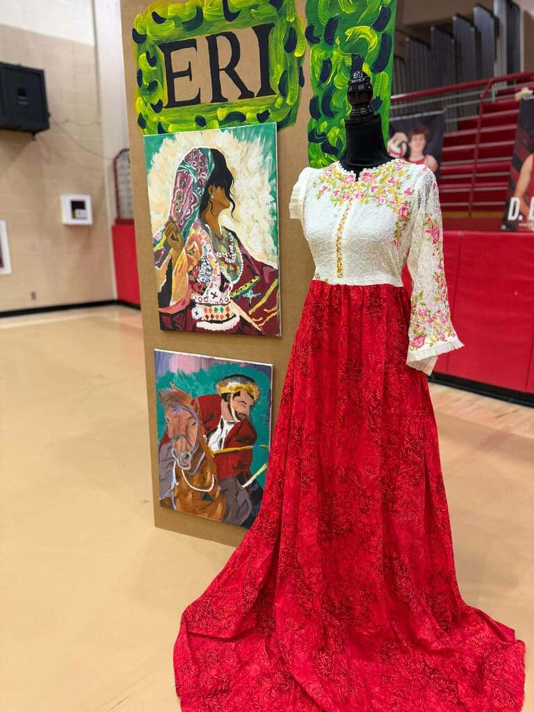 Mannequin wearing a white embroidered top and long red skirt displayed beside student paintings.