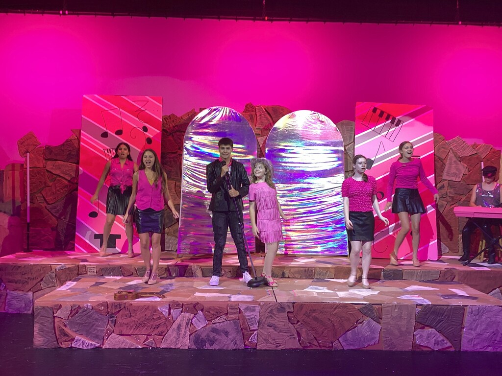 Students rehearse a musical number from High School Musical 2 JR onstage with colorful costumes, props, and set pieces.