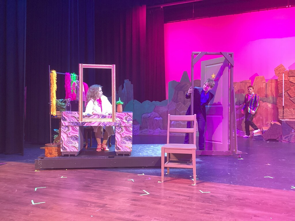 Students rehearse a scene from High School Musical 2 JR onstage with colorful set pieces and costumes.