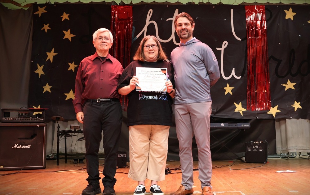 **Alt text:** Three adults stand together on a stage during an awards presentation. The person in the center holds a certificate while posing with two presenters on either side. Behind them is a black backdrop decorated with gold stars, red metallic streamers, and large handwritten-style lettering, creating a celebratory atmosphere. Musical equipment, including amplifiers and a keyboard, is visible on the stage. The group smiles for a recognition photo during what appears to be a staff appreciation or awards event.