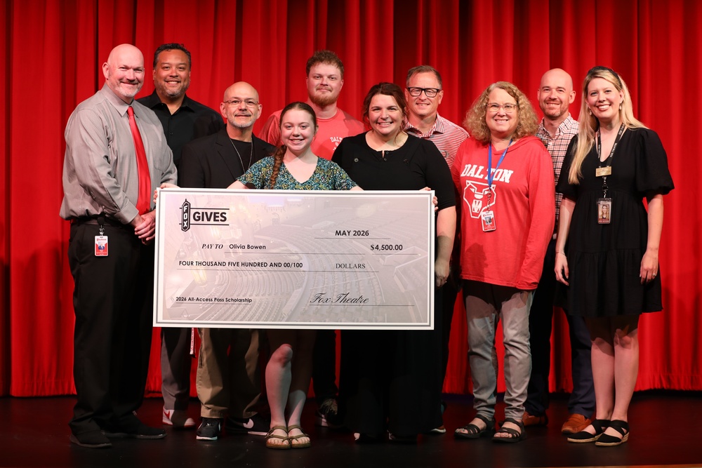 A group of students, staff, and administrators stand on a stage in front of bright red theater curtains while holding an oversized scholarship check. The check, made out to Olivia Bowen for $4,500, is labeled as a 2026 All-Access Pass Scholarship from the Fox Theatre. Everyone is smiling as they pose together for the presentation photo.