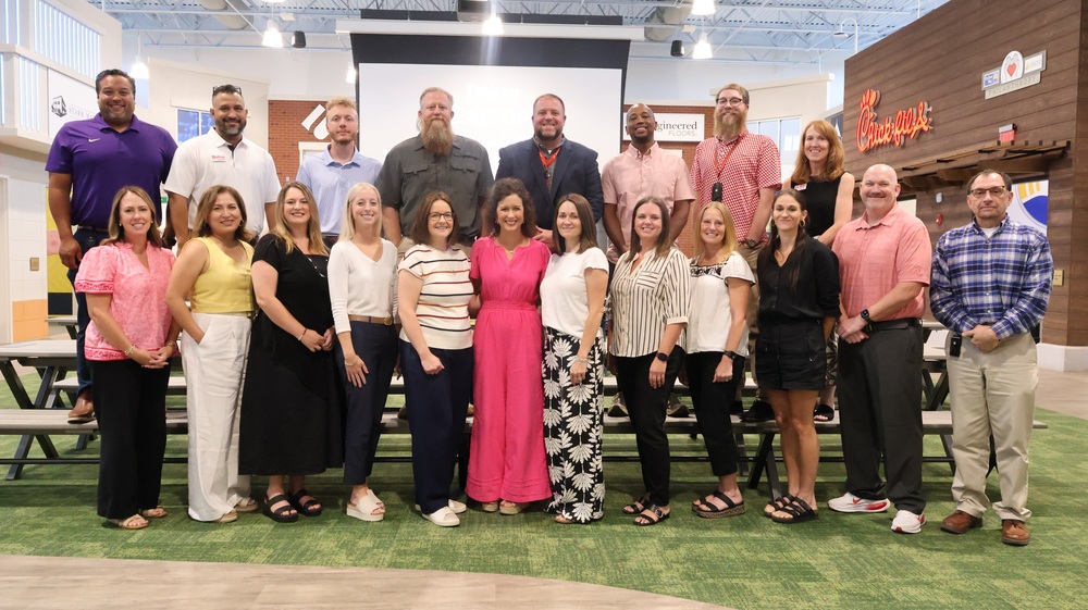 A large group photo of seventeen colleagues lined up in two rows against an indoor backdrop featuring a projection screen and simulated storefronts, including a Chick-fil-A sign. The back row consists of eight men and women in business-casual attire, while the front row features nine women and men standing side by side, including a woman in a bright pink jumpsuit centered in the front.