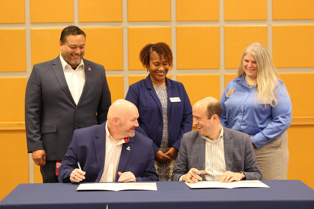Five education leaders smile during a partnership signing ceremony. Two men seated at a table sign documents while three colleagues ā two women and one man ā stand behind them, smiling. The group is dressed in business attire, posed in front of a wall with orange panels.