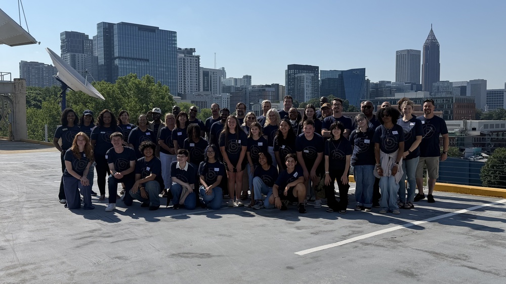 A wide outdoor group photograph showing over thirty students, educators, and chaperones posed on an open concrete rooftop deck on a sunny day. The group is arranged in multiple rows with individuals standing, kneeling, and sitting in the front. A prominent city skyline with modern skyscrapers, including the distinctive Bank of America Plaza tower, forms the background backdrop under a clear blue sky.