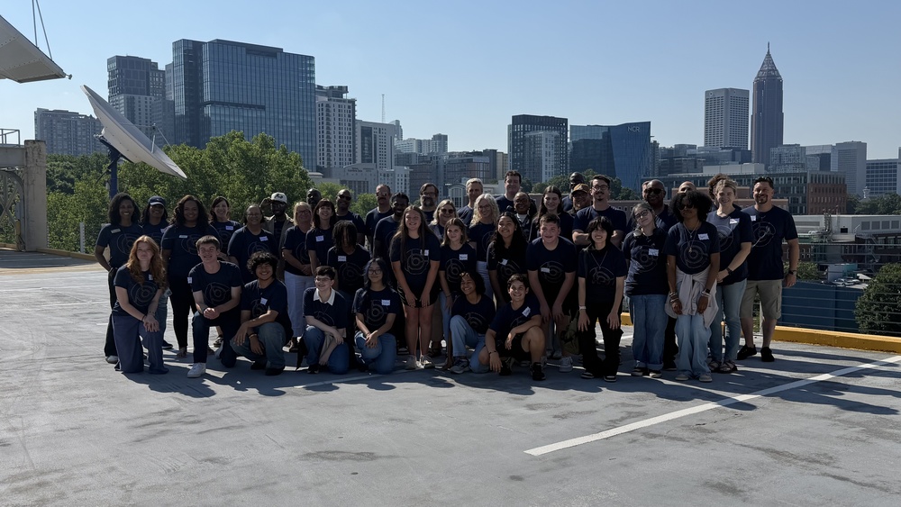A wide outdoor group photograph showing over thirty students, educators, and chaperones posed on an open concrete rooftop deck on a sunny day. The group is arranged in multiple rows with individuals standing, kneeling, and sitting in the front. A prominent city skyline with modern skyscrapers, including the distinctive Bank of America Plaza tower, forms the background backdrop under a clear blue sky.