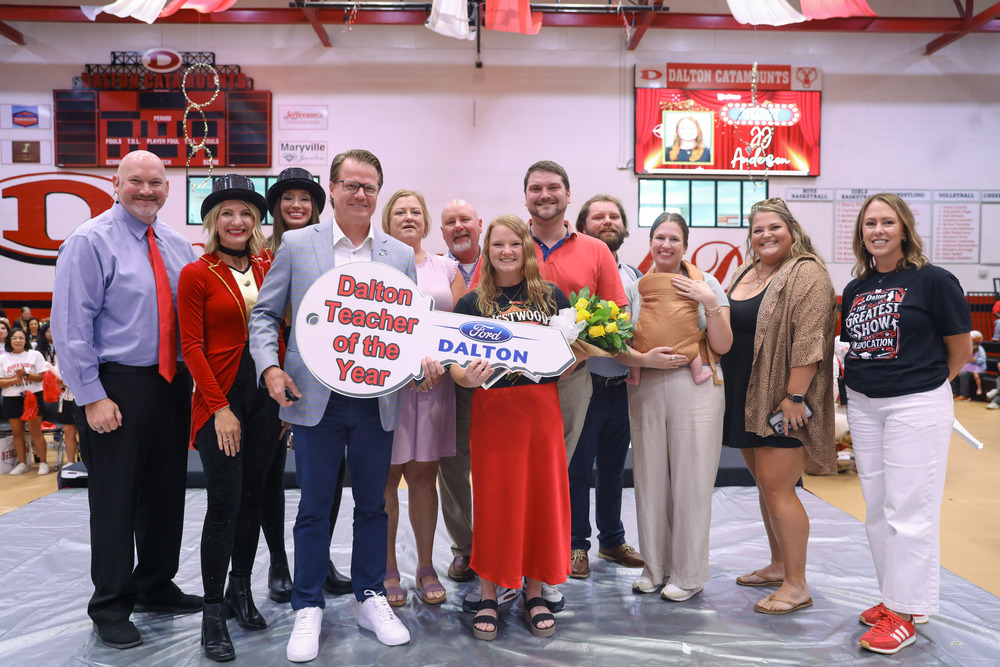 A group of ten people stand together on a stage inside a gymnasium, celebrating the Dalton Teacher of the Year award. In the front center, a man in a light blue jacket and a woman in a red skirt hold a large white novelty key that reads "Dalton Teacher of the Year" with the Ford and Dalton logos. The surrounding group includes school administrators, colleagues, and family members, with digital scoreboard displays visible in the background.