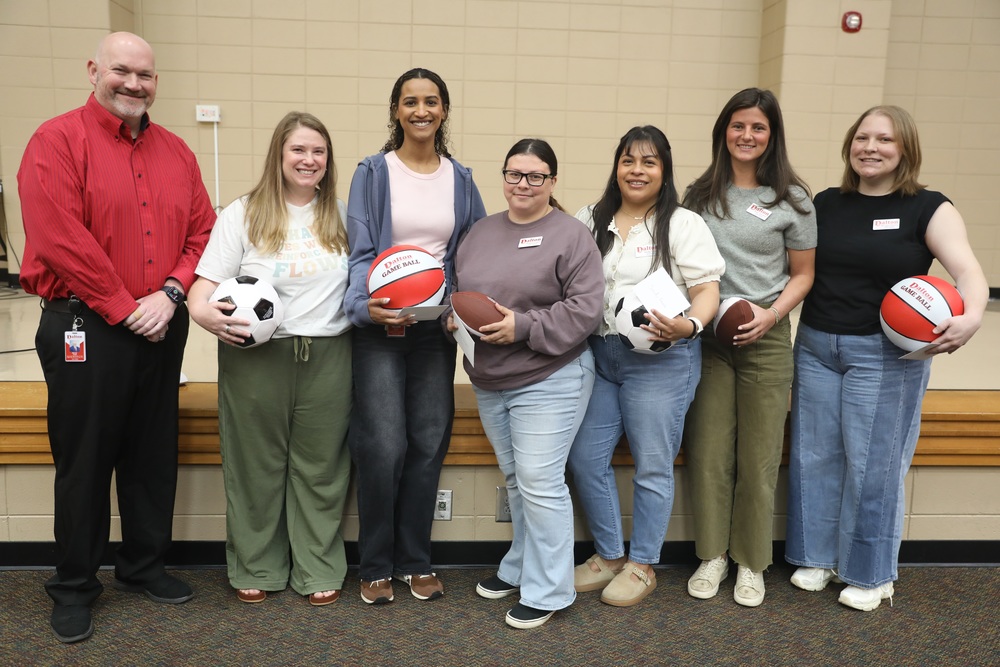 A group of seven adults stands together indoors, smiling at the camera. Several hold sports balls, including soccer balls, footballs, and red game balls, suggesting a school or team-related activity or recognition event.